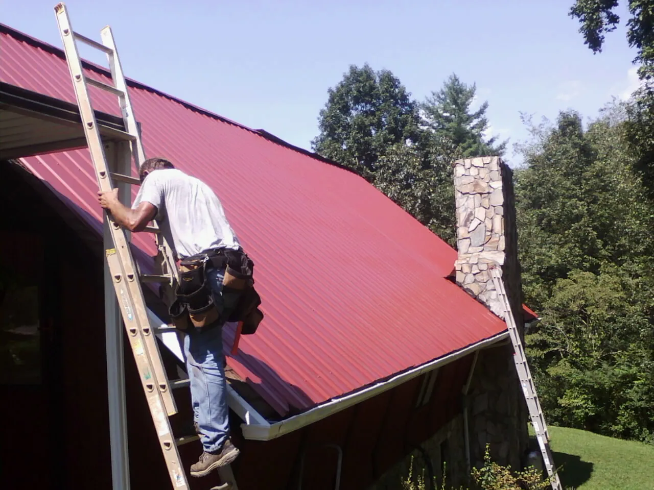 Roofer on a ladder beside a red metal roof and stone chimney — Soffit & Fascia Repair by Roofing Brothers in Port Gamble